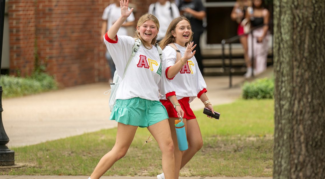 Two sorority students walking and waving at the camera,
