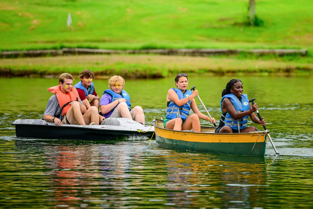 Three male students sit in a paddle boat while two female students row a canoe on the lake at butter and egg adventures.